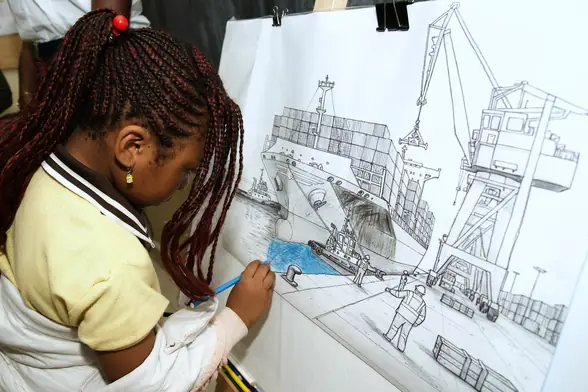 Une élève colorie au crayon un grand dessin de porte-conteneurs à quai